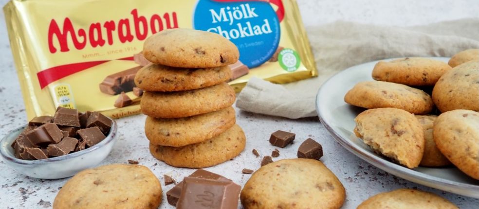 Gestapelte Cookies mit Marabou-Vollmilchschokolade Stückchen, daneben Schokostücke in einer Schale, weitere Cookies auf einem Teller und eine Marabou King Size Vollmilch Tafel in goldener Verpackung