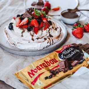 Pavlova mit Sahne, frischen Beeren, Marabou Stückchen und Schokoladensoße, daneben eine geöffnete Tafel Marabou Salzlakrits