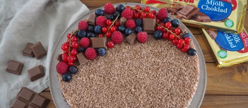No-Bake-Schoko-Cheesecake mit Schokoraspeln, frischen Beeren und Marabou-Schokoladenstücken, serviert auf Teller mit Marabou King Size Tafeln im Hintergrund